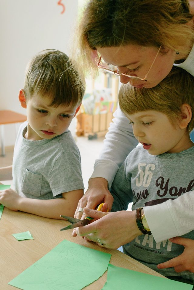 Erwachsene begleitet zwei Kinder beim Schneiden mit der Schere &mdash; Feinmotorik &uuml;ben