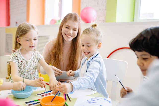 Kinder malen fr&ouml;hlich mit Wasserfarben, eine Betreuerin begleitet sie spielerisch